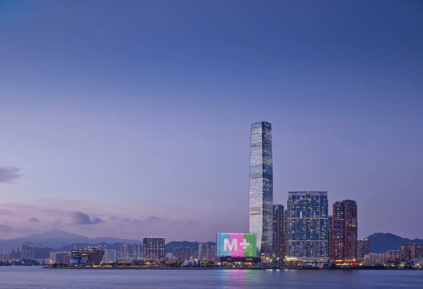 從香港島方向遠眺，看見維多利亞港對岸的M+大樓。相片的右邊是幾幢大樓，包括一座長方形的大樓，上面以三元色投射着「M+」字樣。
